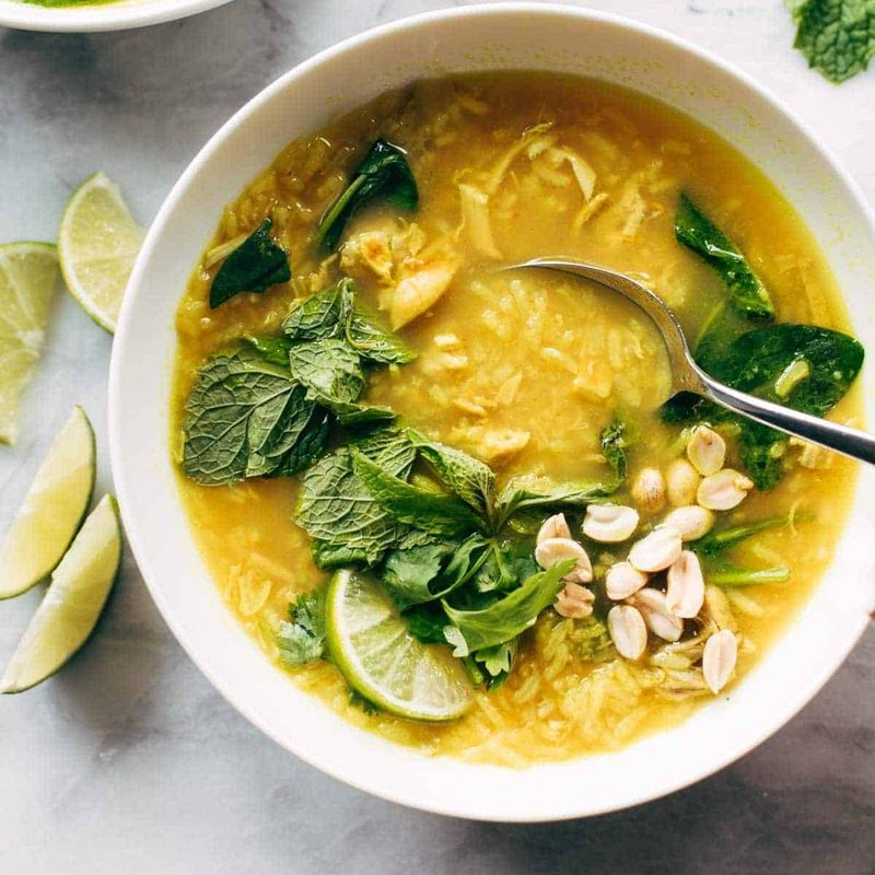 Chicken and rice soup in a bowl with a spoon. There are fresh herbs on top and lime wedges on the side.