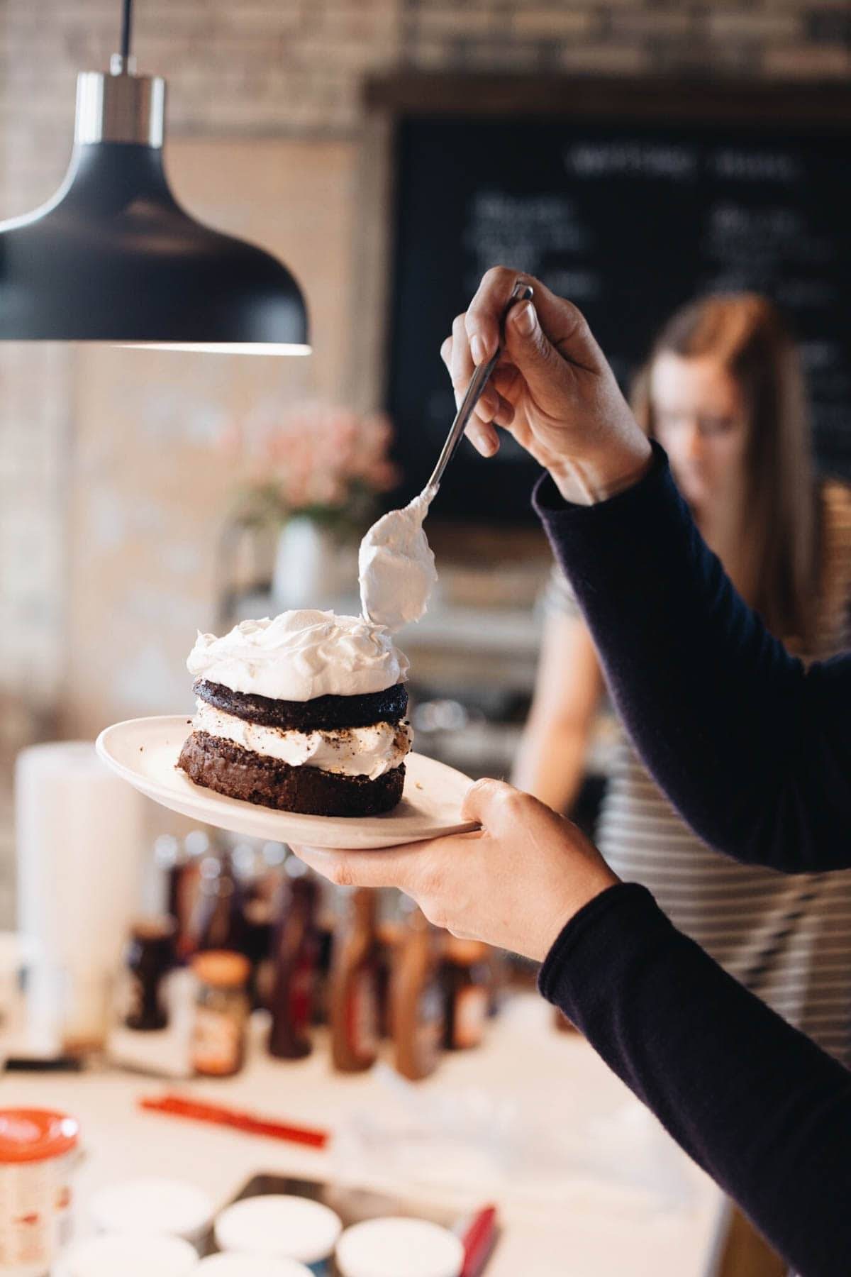 Hand holding a spoon of whipped cream decorating a small chocolate cake.